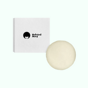 A close-up of a product presentation. 
A small, square, white box with a black graphic of a woman with an afro hairstyle and the words "Natural Moxy" in a stylized, handwritten font. 
Below and slightly offset to the right of the box is a round bar of light beige-cream-colored solid conditioner bar. 
The background is light green. The lighting is even, casting no significant shadows and highlighting the product and the box.
